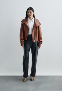 Veste en cuir marron à large col, associée à un pantalon en similicuir noir et des talons dorés pointus. Chemise blanche en dessous.