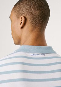 Pruhovaná polo košeľa v svetlomodrej a bielej farbe, so stojacím golierom a vyšitým detailom "LONDON S.1973" na zadnej strane. Texturovaná látka.