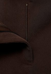 Un tissu marron avec une texture lisse présente une fermeture éclair visible sur le côté droit. Le tissu se plie légèrement, montrant sa souplesse.