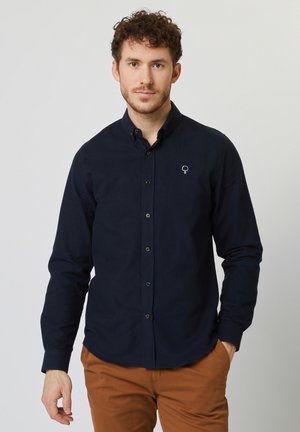 Camisa azul marino de botones con mangas largas, que presenta un logo de árbol blanco en contraste en el pecho. Textura suave, corte relajado.
