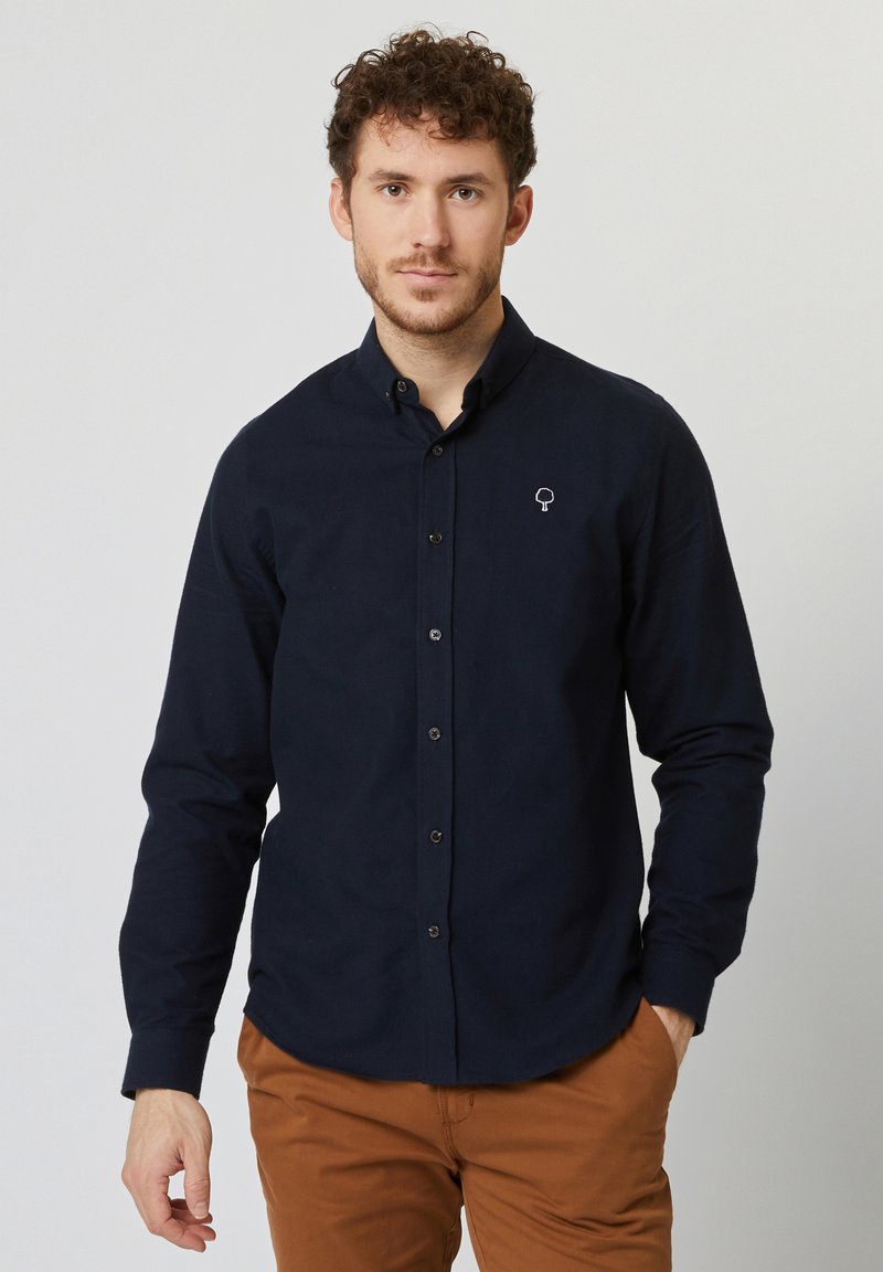 Camisa azul marino de botones con mangas largas, que presenta un logo de árbol blanco en contraste en el pecho. Textura suave, corte relajado.