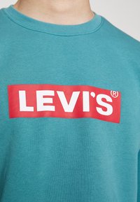 Gros plan sur une chemise bleu sarcelle avec un logo rectangulaire rouge affichant en lettres blanches majuscules le mot "LEVI'S" sur la poitrine.