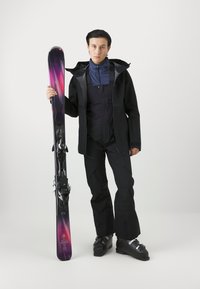 Schwarze Skijacke und -hose mit einer blauen Innenschicht. Der Skifahrer hält einen lila Ski mit einem Gradientmuster und schwarzen Bindungen. Schwarze Skischuhe.