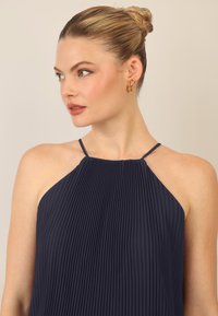 Abito halter plissettato navy con spalline sottili, caratterizzato da righe verticali e una superficie testurizzata. Orecchini con dettagli dorati completano il look.