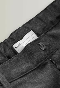 Gråt stof med en struktureret finish, med et mærke med 'NAME' og stiplede linjer, fastgjort med en sort metalbeslag.