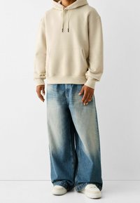 Person som bär en beige hoodie, vida blekblå jeans och vita sneakers, stående mot en ljus, enfärgad bakgrund.