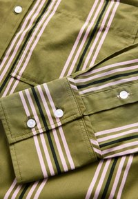 Olijfgroene shirt met roze en donkergroene verticale strepen, voorzien van witte knopen en een enkele borstzak. Smooth katoentextuur.