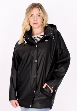 Blonde Frau trägt einen schwarzen Regenmantel mit Kapuze über einem gestreiften Hemd, die Hände in den Taschen, vor einem schlichten weißen Hintergrund.
