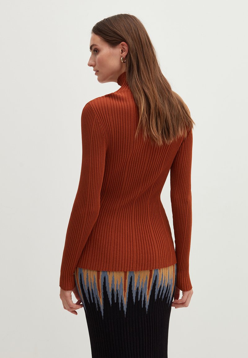 Rippstrickpullover in verbranntem Orange, mit hohem Kragen und langen Ärmeln. Kombiniert mit einem gemusterten Strickrock, der Blau-, Gelb- und Schwarztöne zeigt.