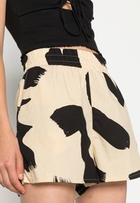 Beige shorts with large black abstract patterns, elastic waistband, and loose fit. Made of lightweight fabric with a soft texture.