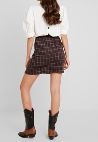 Vrouw in een witte blouse met pofmouwen, een bruine geruite minirok en zwartbruine cowboylaarzen, staande voor een effen grijze achtergrond.