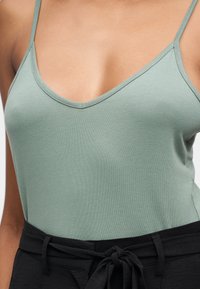 Γυναίκα που φοράει ανοιχτό πράσινο αμάνικο τοπ με λεπτές τιράντες και μαύρο παντελόνι δεμένο στη μέση με υφασμάτινη ζώνη.