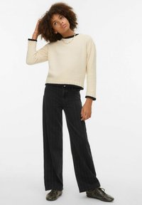 Kremasti džemper s teksturiranim završetkom i crnim rebrastim rubom, upareni s crnim širokim hlačama s prugama i uzorkovanim cipelama.