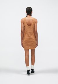 Mežģīņu adījuma, bez piedurknēm kleita siltā brūnā krāsā ar apaļu aizmuguri un vaļēju piegriezumu, valkāta kopā ar melnām augstām kedām un baltām zeķēm.