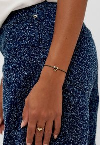 Bracciale nero con charm a forma di cuore dorato, indossato su un polso, su un indumento blu scuro con texture. Design semplice e contrasto di colori.