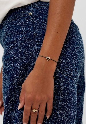 Bracciale nero con charm a forma di cuore dorato, indossato su un polso, su un indumento blu scuro con texture. Design semplice e contrasto di colori.