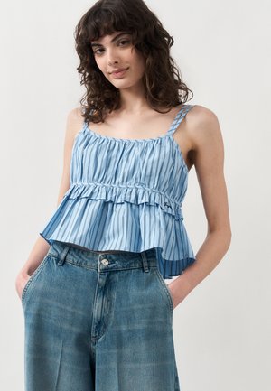 Νεαρή γυναίκα φοράει μπλε ριγέ αμάνικη τοπ με βολάν και ψηλόμεσα φαρδιά μπλε τζιν, με τα χέρια στις τσέπες, χαμογελάει ελαφρά.