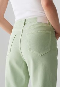 Jeans di denim verde chiaro con vita alta, tasche posteriori e un'etichetta bianca semplice sulla cintura posteriore; superficie liscia e vestibilità sartoriale.