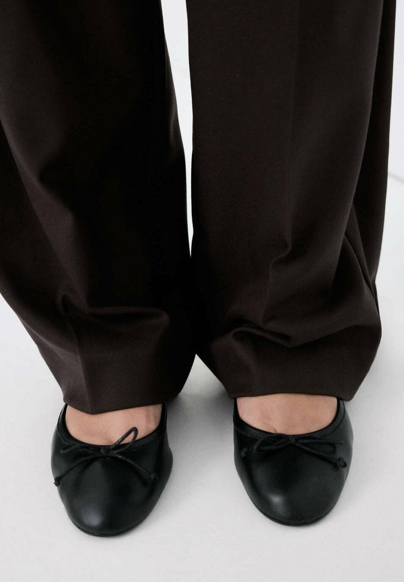 Pieds chaussés de ballerines noires avec petits nœuds, associés à un pantalon ample marron foncé qui couvre les chevilles.
