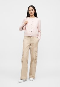 Ροζ ανοιχτόχρωμο ζακέτα με στρογγυλή λαιμόκοψη και κούμπωμα με κουμπιά, σε συνδυασμό με μπεζ παντελόνι cargo φαρδύ με πολλαπλές τσέπες. Λευκά αθλητικά παπούτσια.