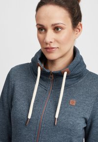 Hoodie i mörkblått tyg med strukturerad yta, med dragkedja, krämfärgade snörningar och bruna läderdetaljer.