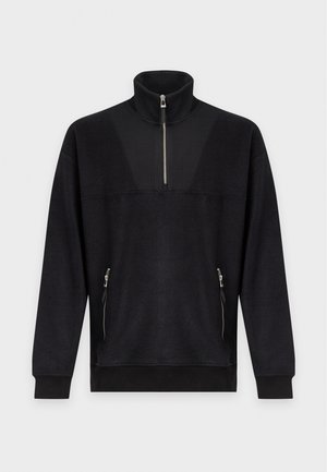 Homme portant un pull zippé noir et un pantalon, tenant un sac cabas en cuir noir, regardant sur le côté sur un fond blanc uni.