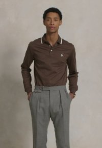 Camisa polo de manga longa castanha com colarinho branco em contraste, combinada com calças ligeiramente cinzentas com pregas e bolsos laterais.