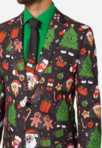 Vīriešu žakete ar krāsainu Ziemassvētku tematisku apdruku, kurā ir koki, dāvanas un svētku varoņi uz melna fona, apvienota ar zaļu kreklu un melnu kaklasaiti.