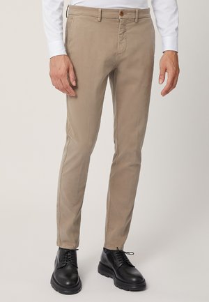 Chinos in cotone beige con vestibilità slim, dotati di chiusura frontale con bottone, passanti per la cintura e tasche laterali, abbinati a scarpe eleganti nere.