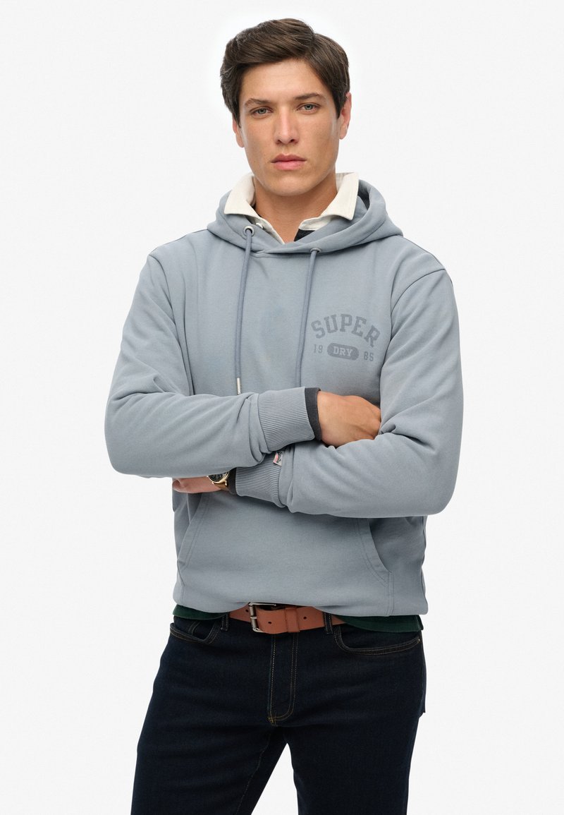 Sudadera gris con capucha, un logo frontal que dice "SUPER" y cordones. Combinada con jeans oscuros y un cinturón marrón, diseño clásico.