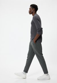 Maglietta a maniche lunghe grigia a costine abbinata a pantaloni tapered grigio scuro. Il modello indossa sneakers bianche, mentre cammina contro uno sfondo minimalista.