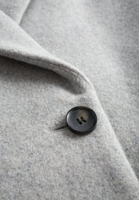 Szara wełniana tkanina o miękkiej fakturze, z dużym czarnym guzikiem na cztery dziurki oraz złożonym kołnierzem. Obecne subtelne tonalne odcienie.