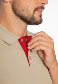 Béžová polo košile s červeným akcentem uvnitř límce, s knoflíkovým zapínáním a texturovanou tkaninou, částečně rozepnutá u výstřihu.