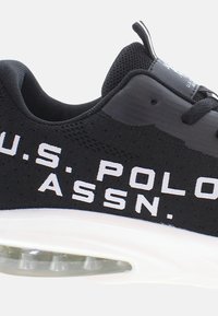 Fekete sportcipő, mely hálós szövetből készült, texturált felülettel. Jellemzője a kiemelkedő fehér márkázás, párnázott talp és egy látható légzóna.