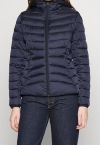 Mørkeblå quilted jakke med frontlynlås, båret over mørke denimjeans, vist fra skuldrene til hoften.