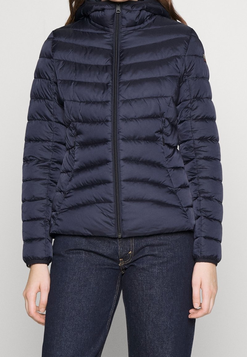 Mørkeblå quilted jakke med frontlynlås, båret over mørke denimjeans, vist fra skuldrene til hoften.