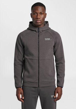 Homme debout portant un sweat à capuche zippé gris foncé et un pantalon de survêtement assorti avec de petits logos blancs hummel sur la poitrine et la cuisse.
