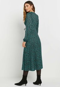 Robe teal à manches longues avec un imprimé léopard noir, présentant une taille cintrée et une jupe fluide. Associée à des bottines noires.