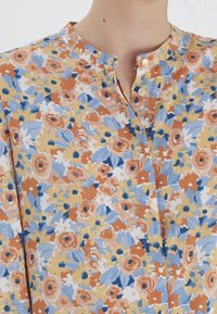 Chemise florale en mélange de coton présentant des motifs orange, jaune et bleu, avec un col rond et une fermeture à boutons. Tissu texturé avec une finition douce.