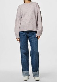 Mujer vistiendo un suéter suelto de color rosa pálido con motas, unos vaqueros de pierna ancha y puños de color azul, y zapatillas blancas, contra un fondo gris liso.