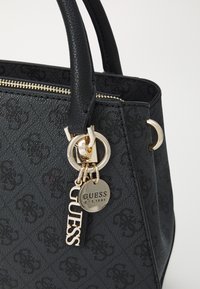 Black textured tote bag with dual handles and gold-tone hardware. Features a zipper closure and branded charm with "GUESS" inscription.