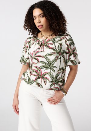 Femme aux cheveux bouclés portant un chemisier blanc à motif de feuilles de palmier vertes et roses et un pantalon blanc, posant la main sur la hanche.