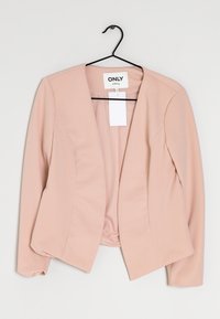 Lichtroze blazer met ribbeltextuur, open ontwerp en een label eraan vast. Geen knopen of sluitingen zichtbaar. Hangt aan een zwarte haak.