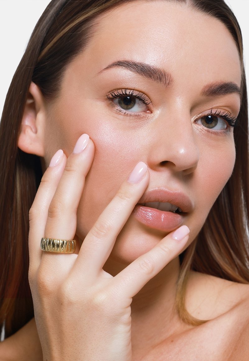 Gouden ring met een verticale ribbelstructuur, gedragen aan een hand met gemanicuurde lichtroze nagels, tegen een neutrale achtergrond. Glad huid en duidelijke gelaatskenmerken.