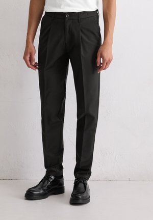 Schwarze maßgeschneiderte Hosen mit flatem Vorderteil, ausgestattet mit einem elastischen Bund und einem glatten, leichten Stoff. Kombiniert mit schwarzen eleganten Schuhen.