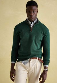 Zaļš džemperis ar ceturtdaļas rāvējslēdzēju, ar ribotām piedurknēm, valkāts virs zilas un baltas rūtainas krekla. Gaiši krāsoti korduroja bikses un pītā josta.