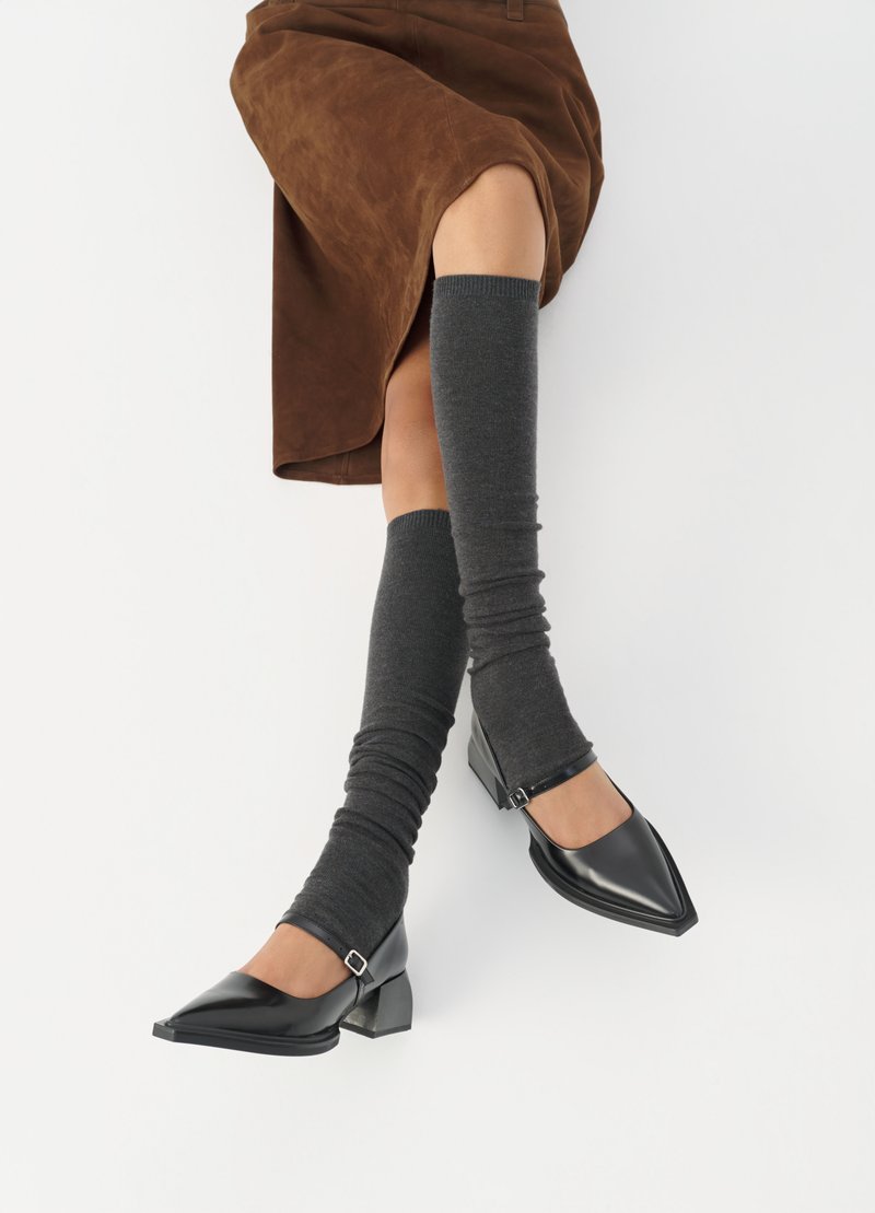 Schwarze lacklederne spitze Schuhe mit Blockabsatz und Knöchelriemen, kombiniert mit grauen gerippten Beinstulpen und einem braunen Wildlederrock.