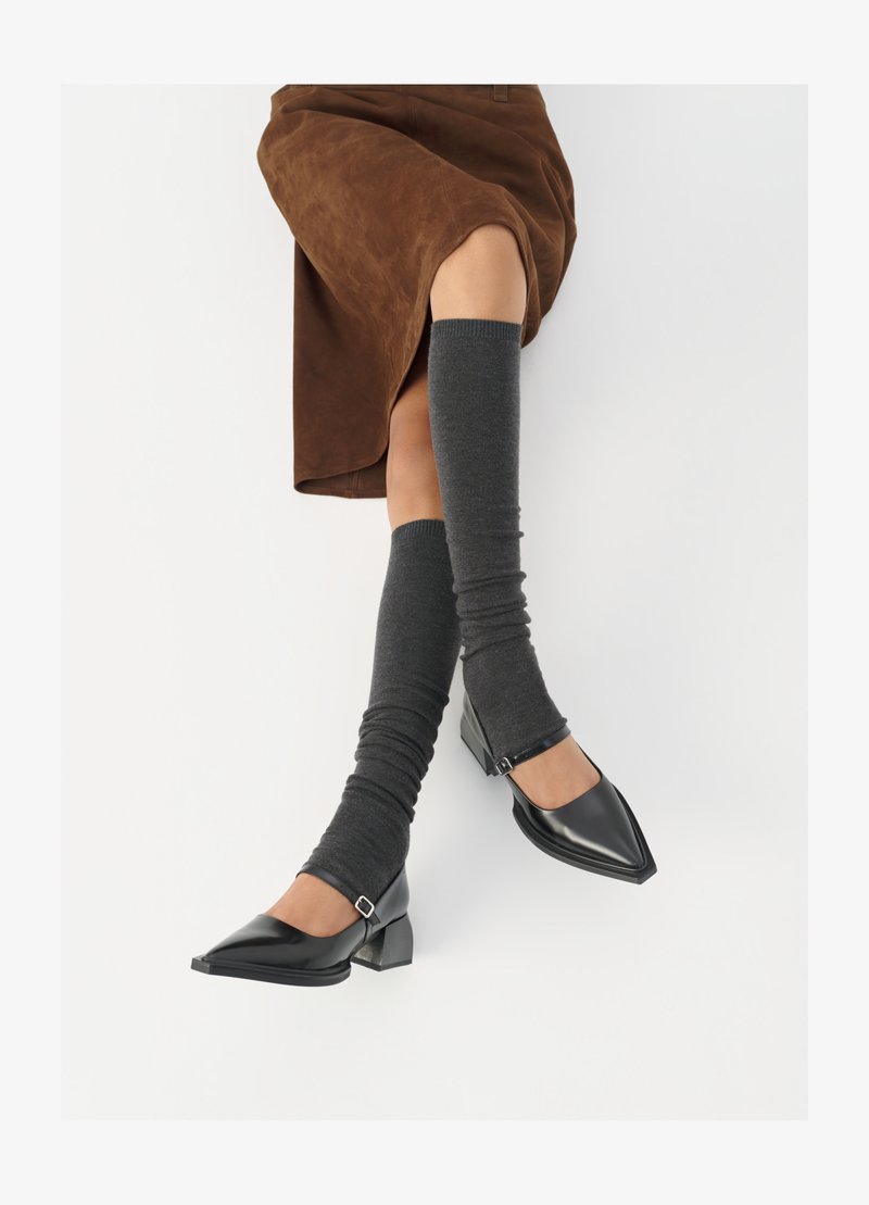 Schwarze lacklederne spitze Schuhe mit Blockabsatz und Knöchelriemen, kombiniert mit grauen gerippten Beinstulpen und einem braunen Wildlederrock.