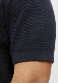 Tamsiai mėlynas trumpomis rankovėmis megztas marškinėliai su tekstūruota raštų ir išilgai megztomis rankogaliais, pasižymintys tarsu prigludusia siluetu ir lygia medžiaga.