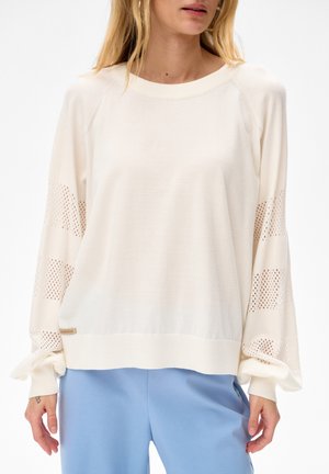 Vrouw draagt een crèmekleurige trui met geperforeerde ballonmouwen en lichtblauwe broek, gefotografeerd van nek tot heupen tegen een witte achtergrond.